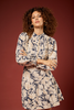 A woman with curly brown hair wearing a beige and navy floral print dress, standing against a solid dark red background.