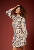 A woman with curly brown hair models a long-sleeve mini dress featuring a beige and navy blue abstract floral pattern, posed against a solid dark red studio background.