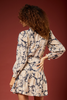 A woman with curly brown hair is shown from the back wearing a beige and navy blue abstract print mini dress with long balloon sleeves, set against a solid dark red background.
