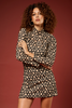 A woman with curly brown hair models a black long-sleeve mini dress with a beige floral pattern against a solid dark red background.
