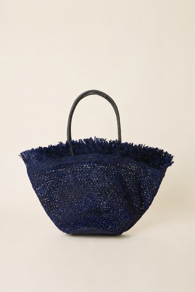 A navy blue woven straw tote bag with a frayed fringe top and a single black handle, photographed against a solid light beige background.